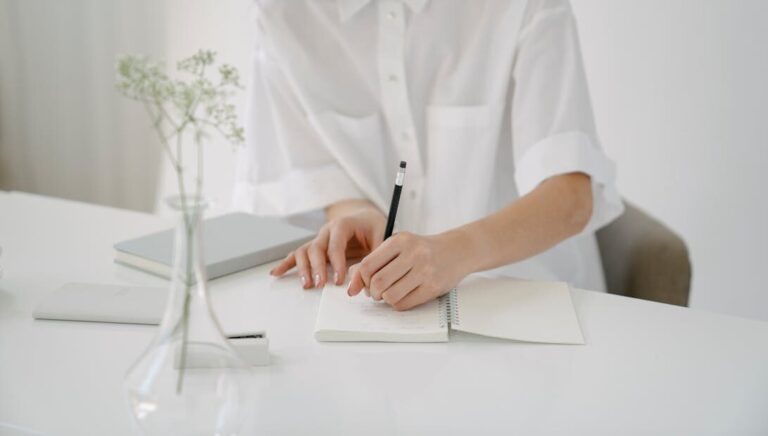 A woman writing in a notebook at a minimalist desk with stationery items and a vase.