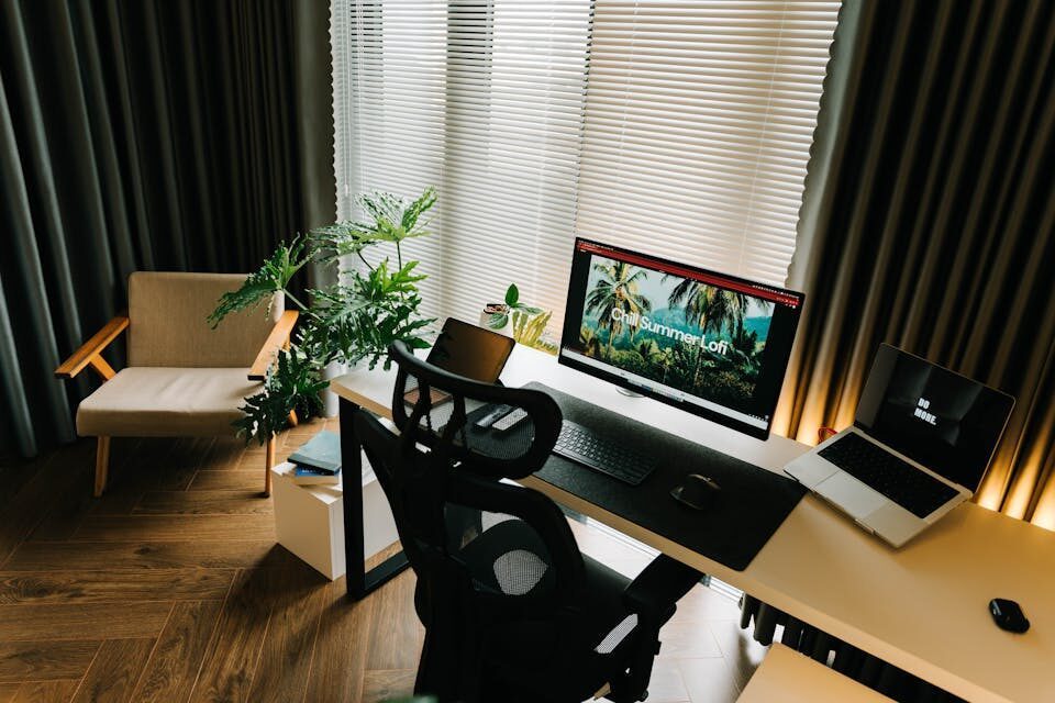A stylish home office setup with a computer, laptop, and indoor plants by a window.