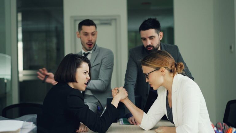 Women in business attire arm wrestling while colleagues look on in surprise.